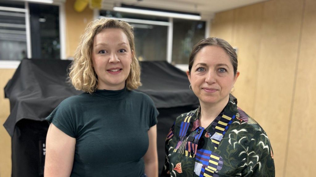 Woman on the left wears a black short sleeved top, has bobbed blonde hair and the woman next to her has a patterned dark blouse and dark brown hair tied back from her face. Behind them are light brown walls.
