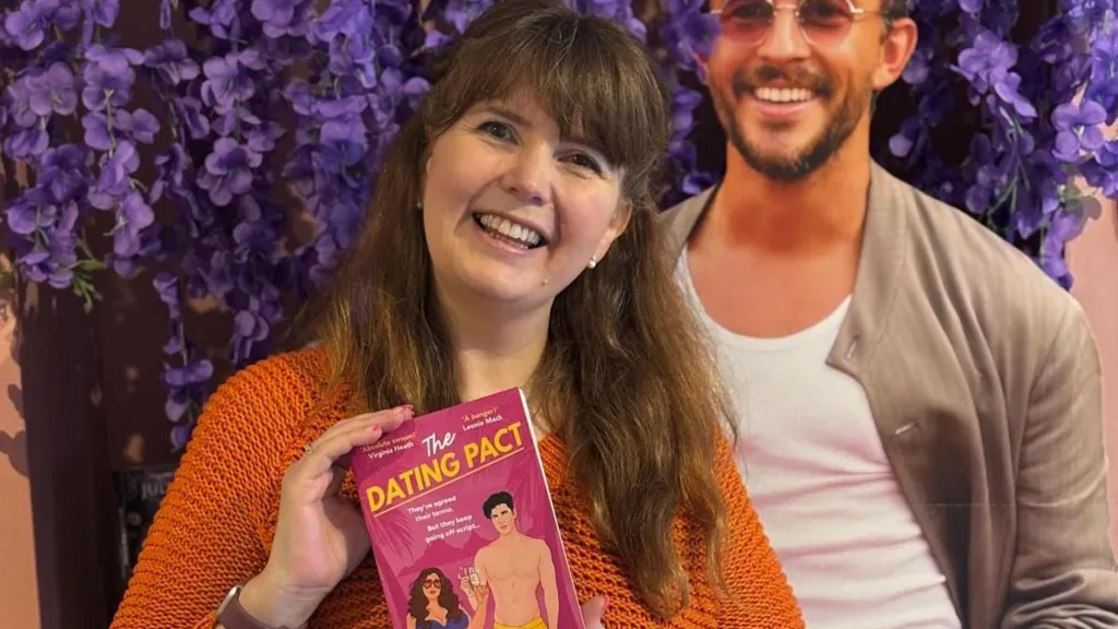 A woman with an orange cardigan on is smiling while holding her book 