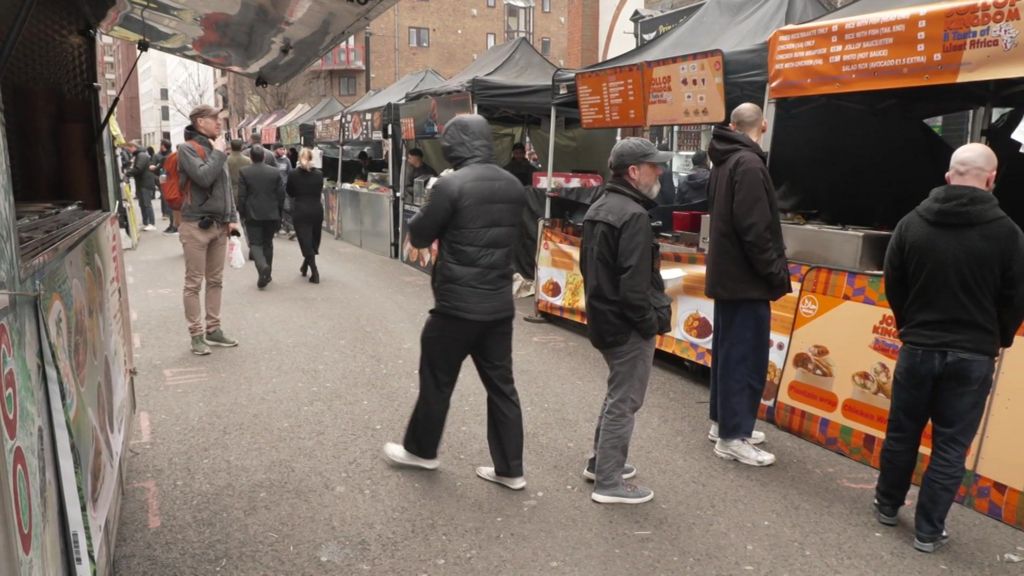 A row of street food stalls with black canopy tents set up along a city street. Several customers queue at the counters while others walk past. The stalls display bright orange menu boards and food images, and apartment buildings can be seen in the background