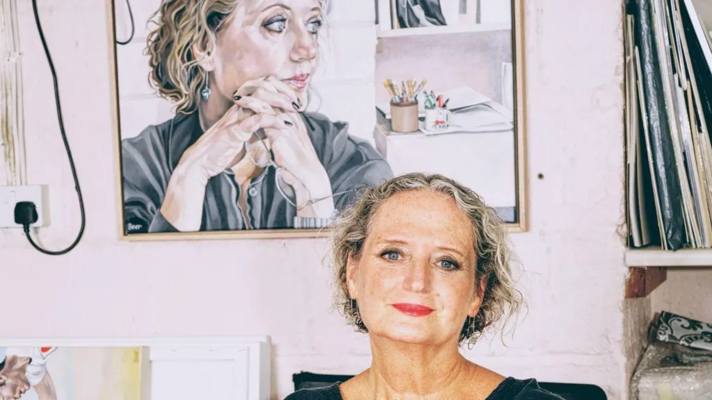 Artist Jo Beer is pictured in front of a self-portrait on the wall behind her.
