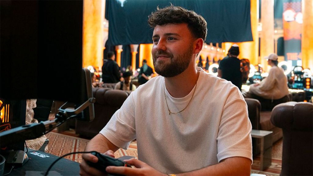 OllMS, a male CoD content creator, wearing a white tshirt while playing on a games console holding a black controller and looking into a screen while smiling. Behind him are several other gamers sitting on sofas.