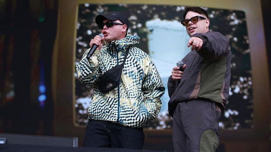 Kneecap performing live at Glastonbury, wearing tracksuits and holding microphones