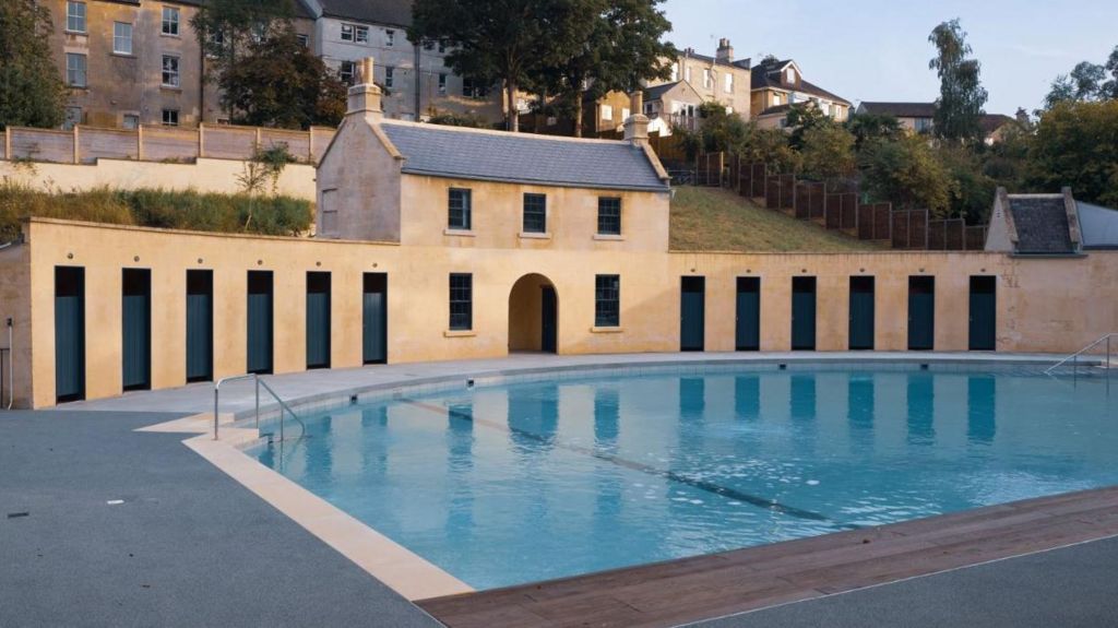 An outdoor pool with no one in it, behind the pool are changing rooms in a semi circle - made from traditional Bath stone