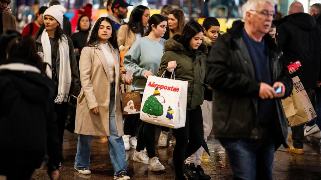 A damp sidewalk in New York City being crossed by men and women carrying shopping bags.