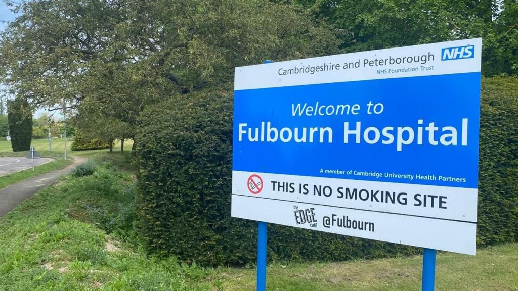 A blue and white sign saying Cambridgeshire and Peterborough NHS Foundation Trust, and Welcome to Fulbourn Hospital, standing on blue legs, against a dark green hedge, on a lighter green verge beside a road