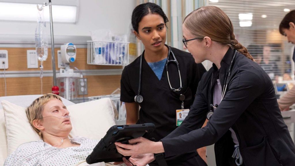 Unknown actor lying in hospital bed with Dr Samira (Supriya Ganesh, centre) and Dr Mel (Taylor Dearden, right) at her bedside showing her a screen on a tablet
