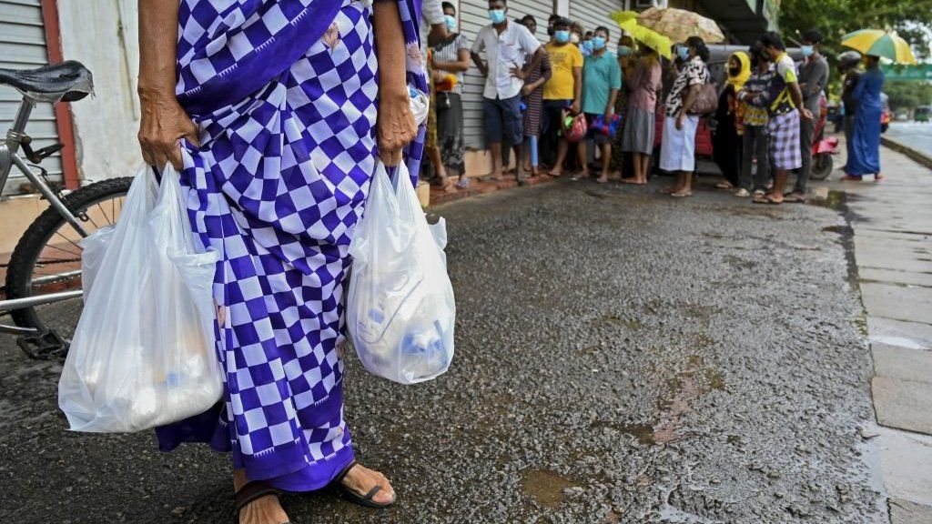 Woman carrying food bags in Colombo while people wait in queue outside a state-run supermarket