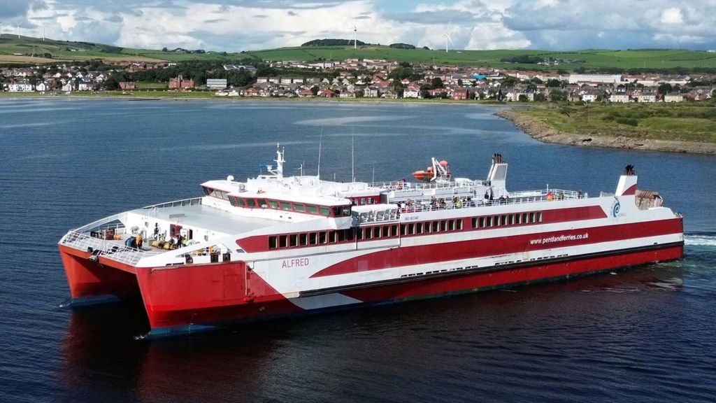 A red and white large catamaran with the name Alfred on the side, pictured in the foreground with a small seaside town in the background.