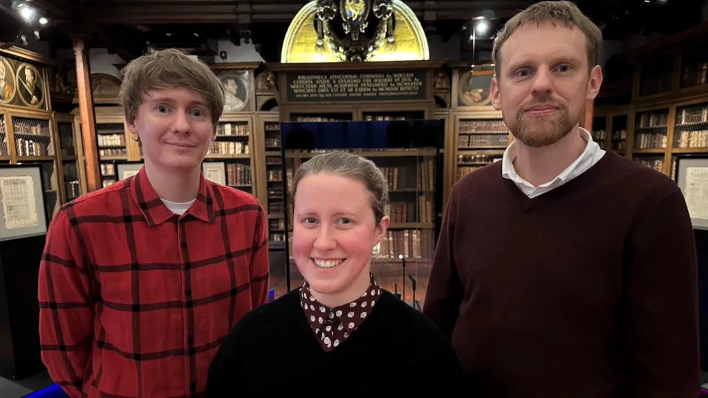 David, Lauren and Tony stand in an ornate old library surrounded by shelves of antique books. David is clean shaven with longish fair hair swept across his forehead. Lauren is smiling with fair hair tied back. Tony has a fair beard and short hair.

