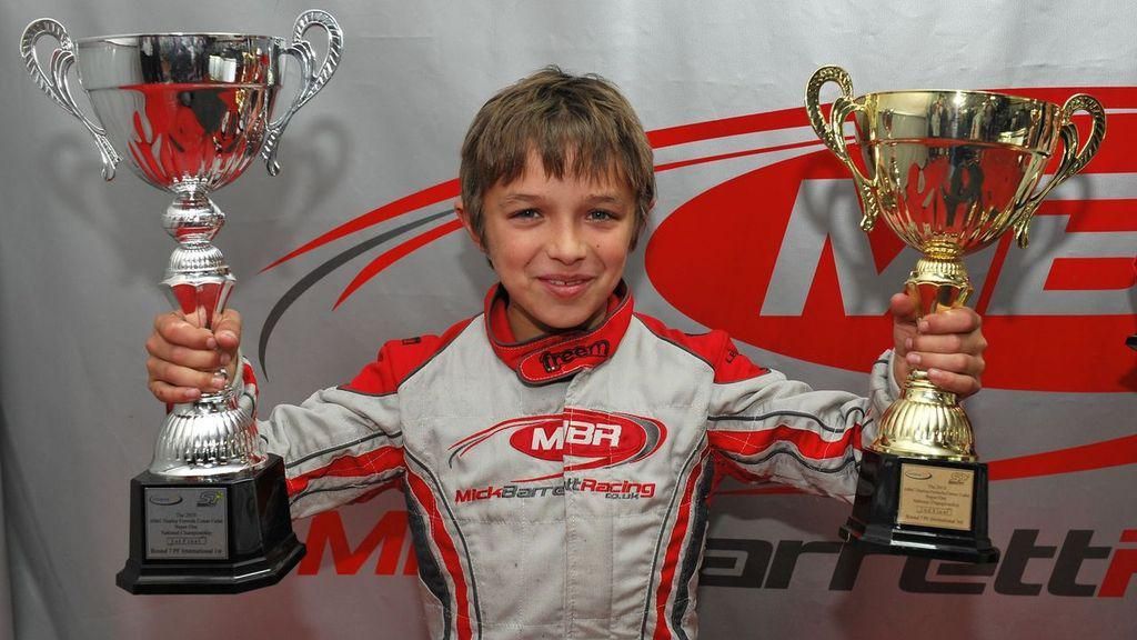 Childhood photo of Lando Norris. He has brown short hair and is wearing a racing suit. He is holding two trophies - one silver, and one gold