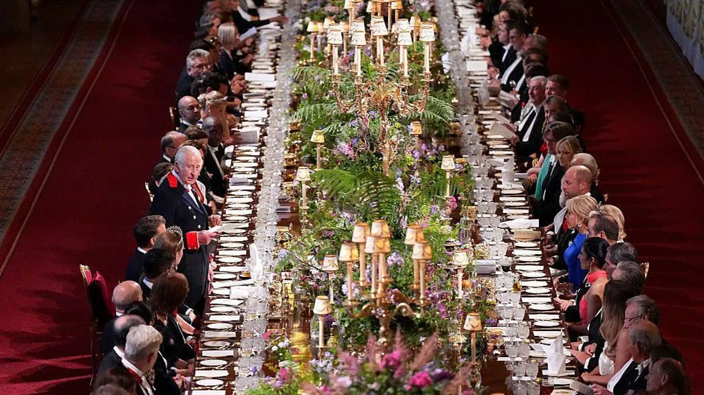 King Charles addresses the banquet's guests, who line a long table covered in flowers, lamps and table-wear.