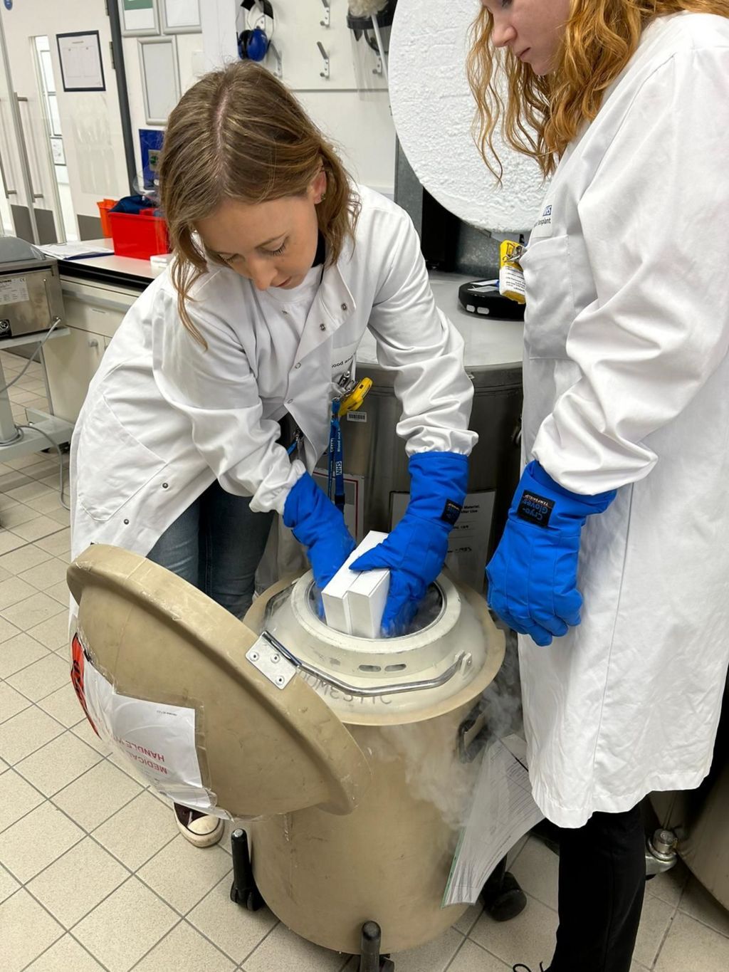 NHSBT staff packing the frozen cord blood cells for transport to Gunner. The two women workers wear white laboratory coats and blue gloves and are putting boxes into the special storage container. 