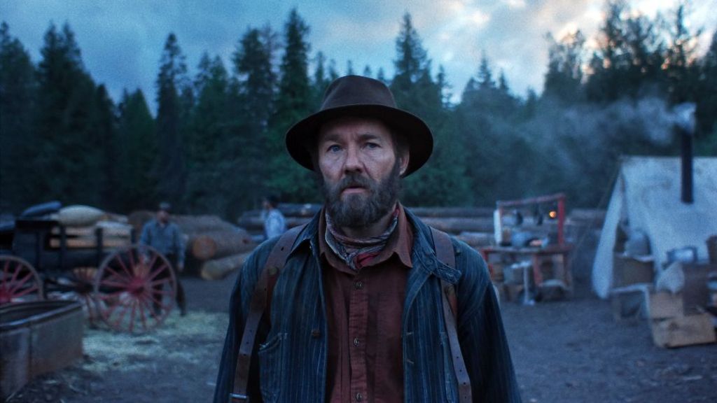 Joel Edgertonin Train Dreams, wearing a hat and looking ahead, with trees in the background