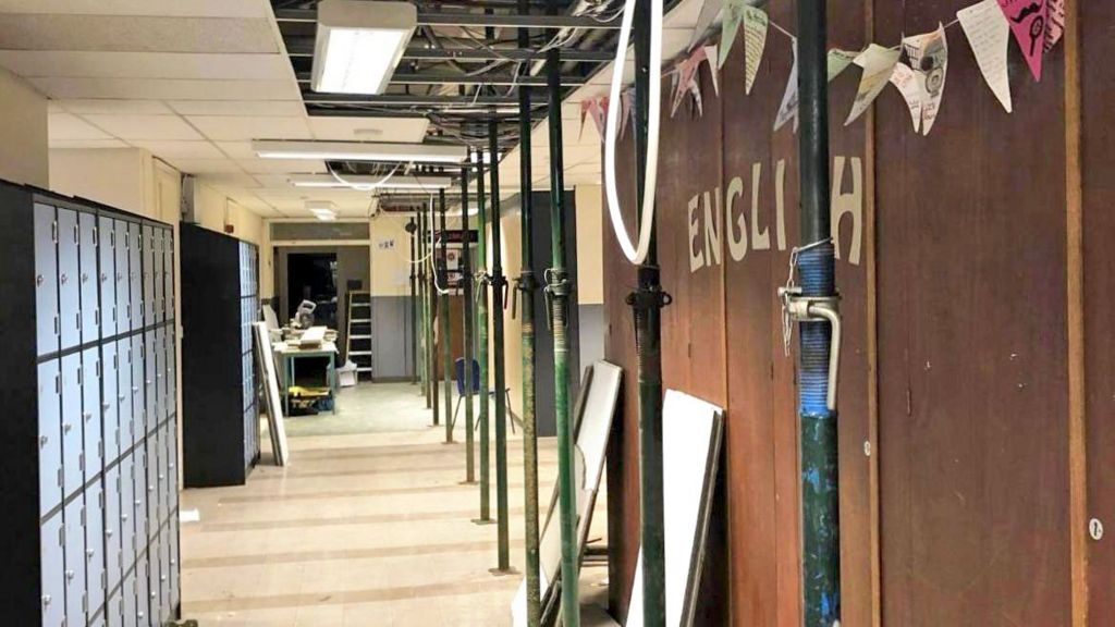 A school corridor with 'English' written on one wall and lockers lining the other. Ceiling panels have been removed and metal poles prop up the ceiling.