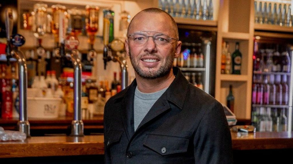 Man with short hair dark hair and a beard, wearing glasses, a grey t-shirt and dark jacket stands in front of a bar