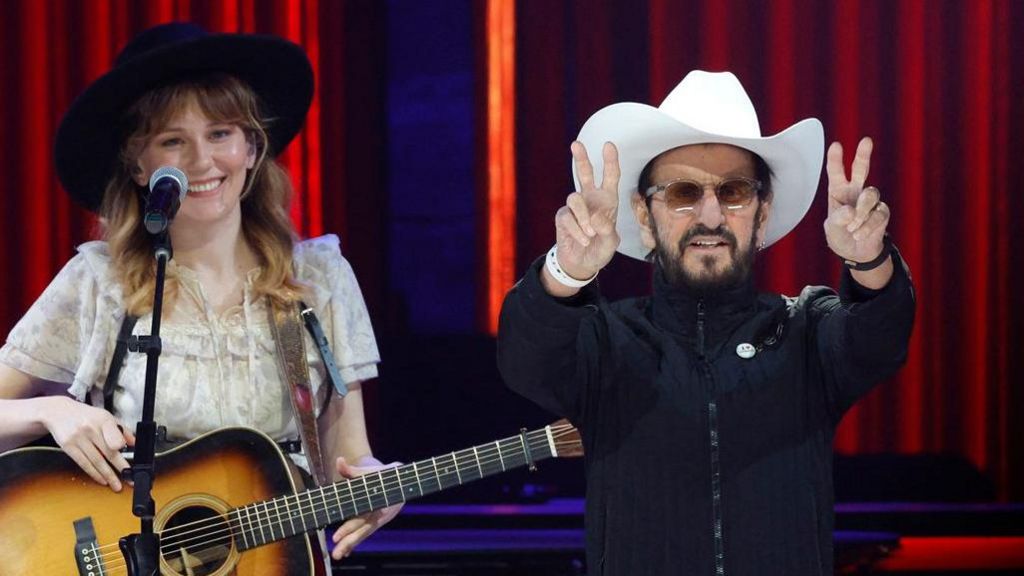 Molly Tuttle and Ringo Starr performed at The Grand Ole Opry in Nashville, Tennessee last year. Molly is wearing a cream-coloured blouse and black hat while holding a guitar. Star has on a white cowboy hat and a black jacket and is doing the peace sign.