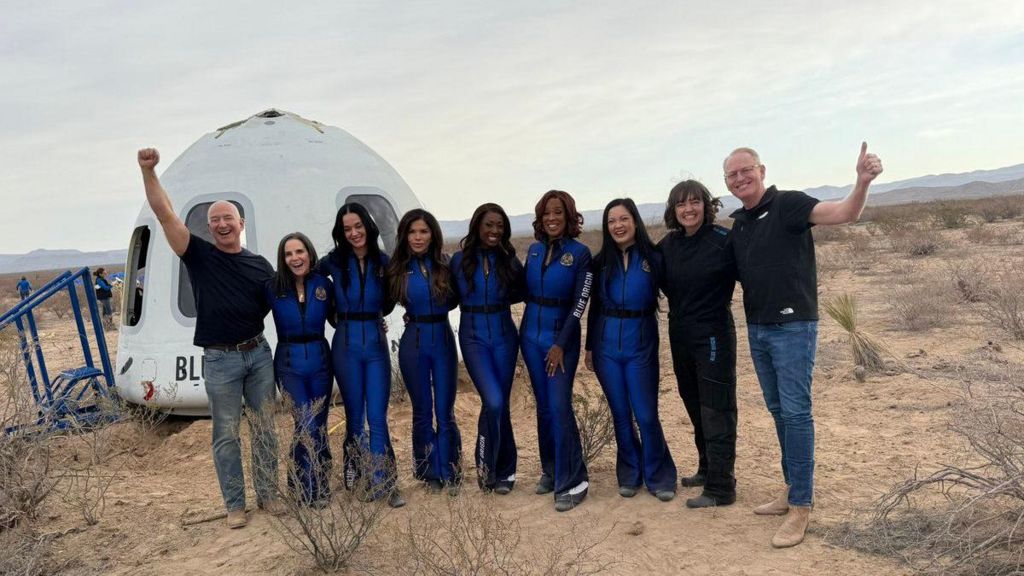 A group of nine people stands in front of the Blue Origin New Shepard capsule in a desert landscape. The six female NS-31 crew members wear matching blue flight suits.
Left to Right: Jeff Bezos, Kerrianne Flynn, Katy Perry, Lauren Sánchez, Aisha Bowe, Gayle King, Amanda Nguyen, Audrey Powers and Dave Limp.
The mood is joyful as the group poses after the successful suborbital spaceflight.