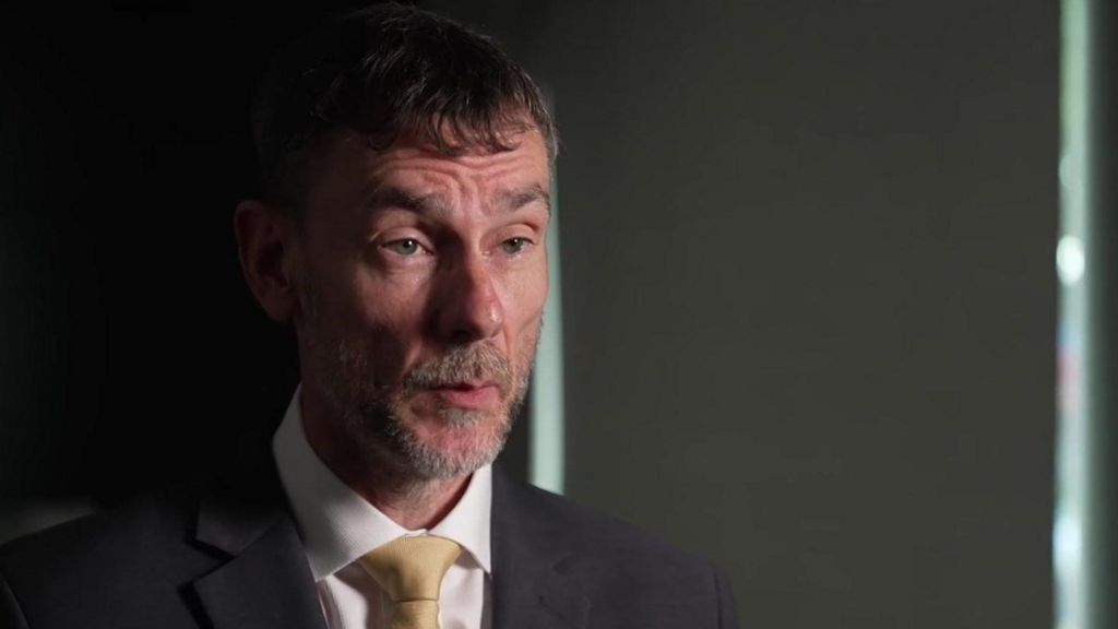 A man in a black suite and golden tie speaks in a dark room with a serious expression. 