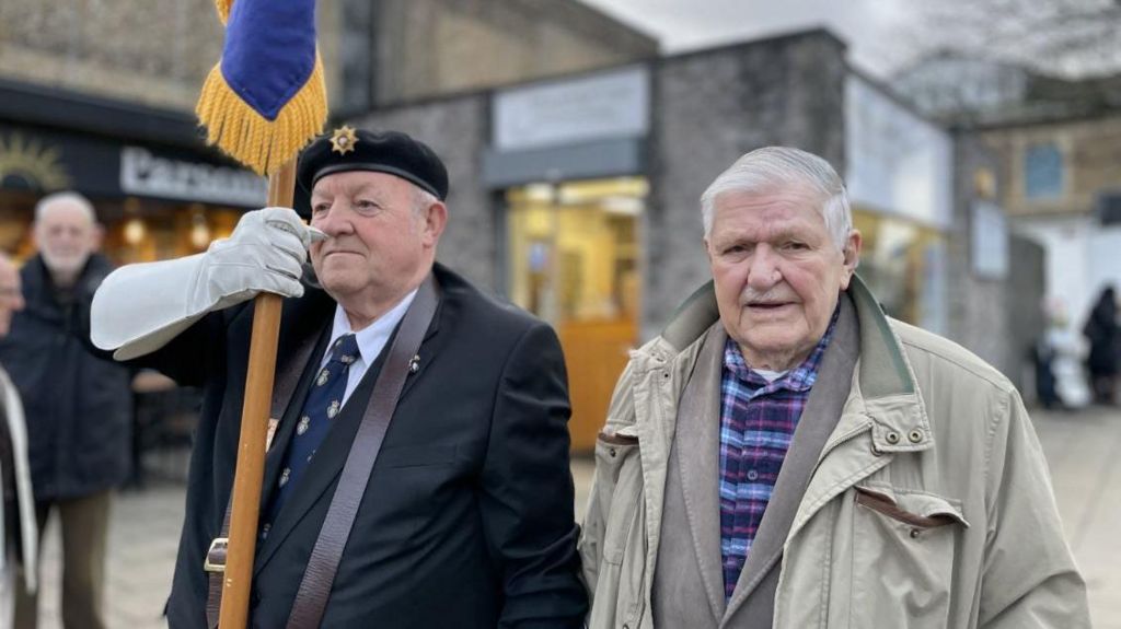 Arthur in a pale green coat looking at the camera, next to a RBL member holding a flag