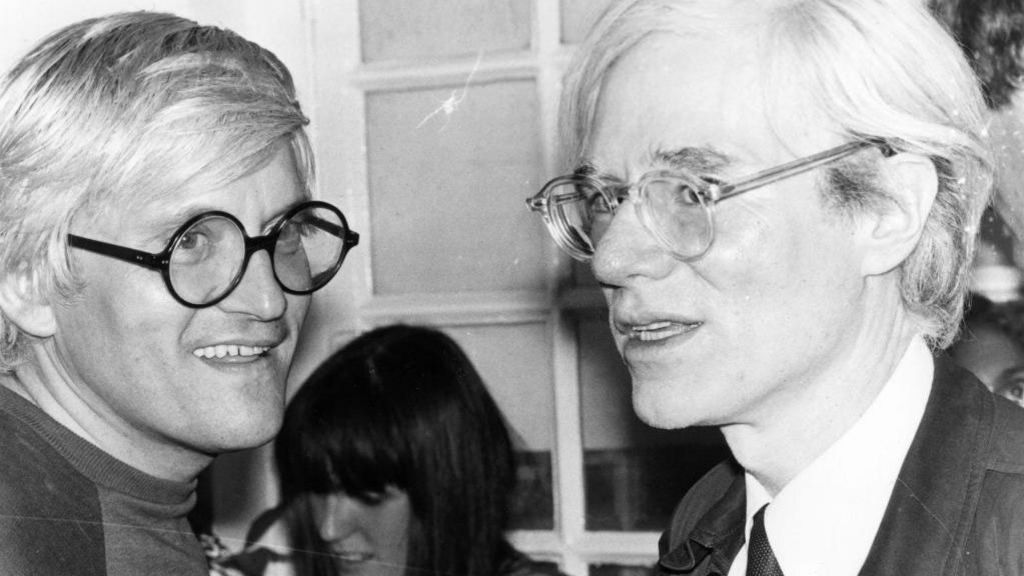 David Hockney and Andy Warhol in the 1970s. They look like mirror images with their white hair and round glasses.
