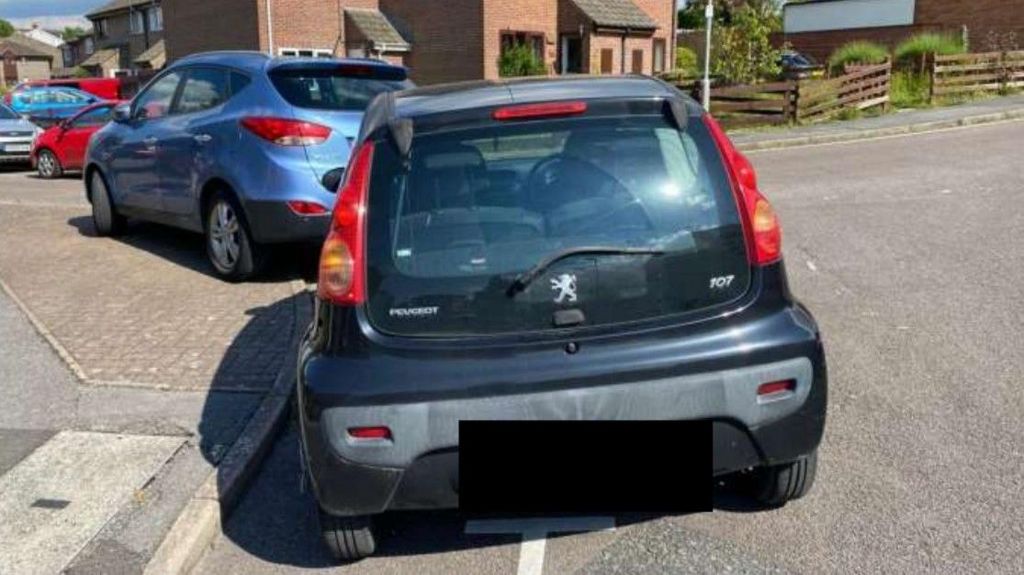 A small black Peugeot car with its numberplate blanked out is parked partly over a white line on the road.