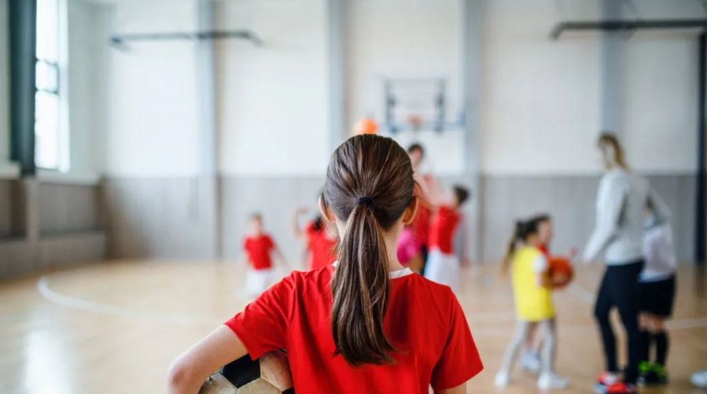 A young girl with a football under her arm facing away from the camera with a PE lesson going on in the background