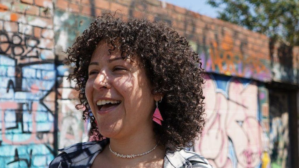 Engy Elboreini smiles at the camera. She has curly dark-brown hair and pink earrings. She is standing in front of a wall spray-painted with graffiti.