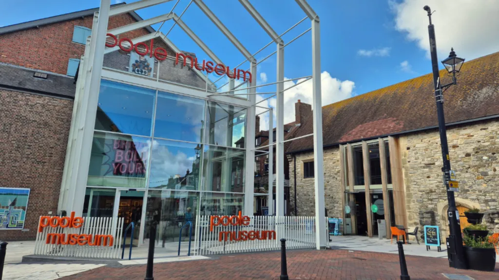 A brick building has a large glass entry. A white fence is out the front of the building and has an orange sign on each fence which reads 'poole museum' in orange letters. 