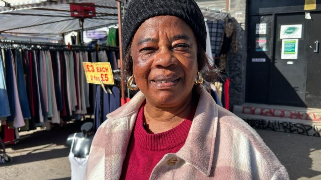 Woman in a pink and white check jacket and black hat smiles at the camera with her clothes stall behind her.
