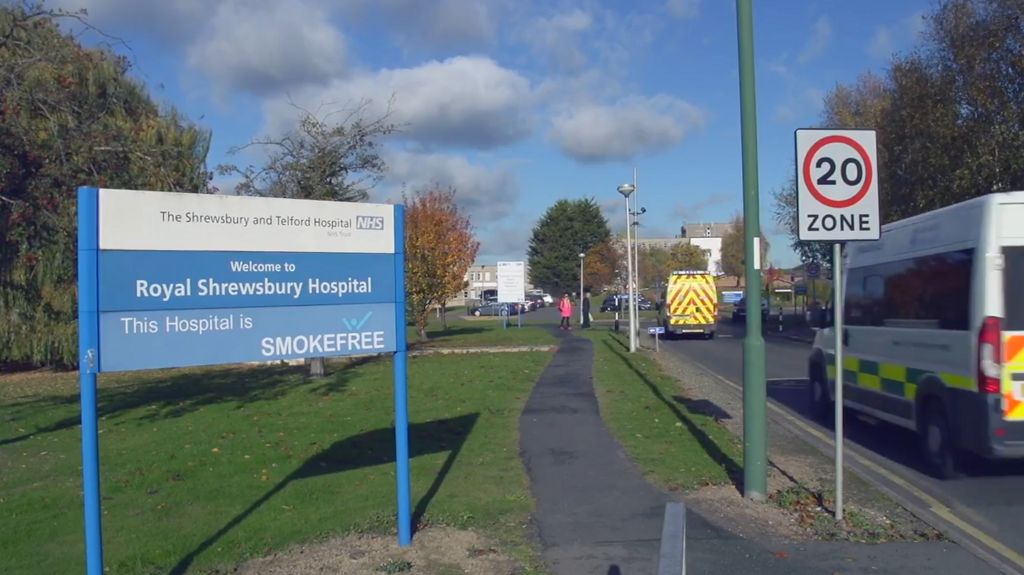 A sign saying "Royal Shrewsbury Hospital" next to a road with a couple of ambulances driving past