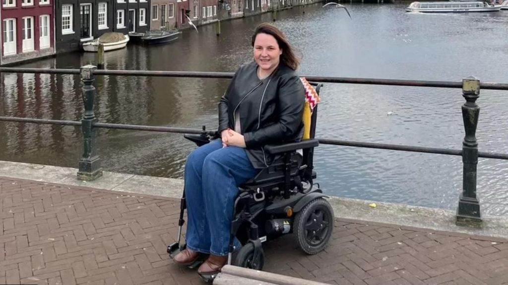 Sam Jennings, who has long brown hair, wears brown boots, jeans and a black leather jacket. She is in a powerchair, with a canal behind her