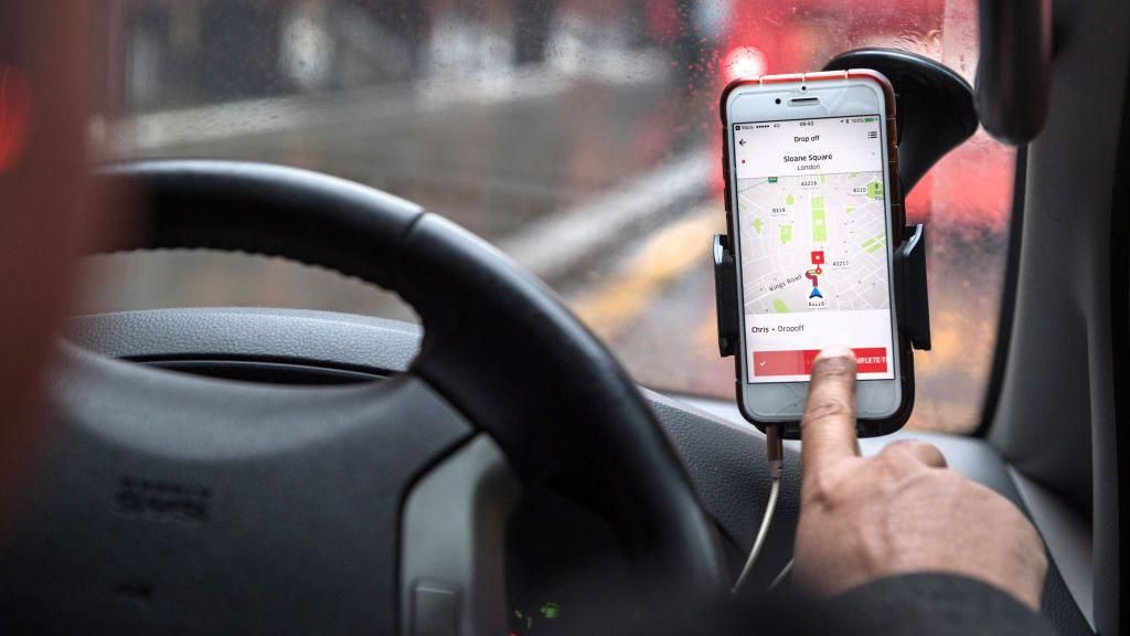 A smartphone mounted on a car dashboard displays a ride-hailing app map, with a driver’s hand reaching towards the screen, seen from inside the vehicle on a wet London street.