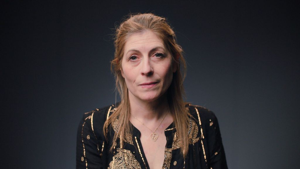 A woman with long light-brown reddish hair is looking straight ahead. She is wearing a black top, with golden threads. She is wearing a Star of David, set in a heart, around her neck