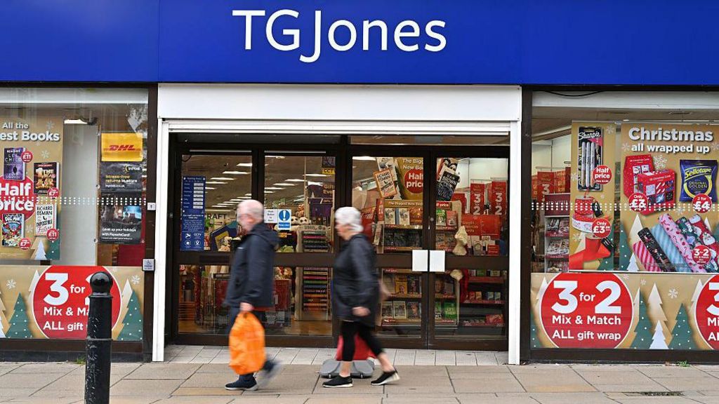 A man and a woman walk past a doorway into a bookshop, with a blue banner sign above the door which says 'TG JONES'.