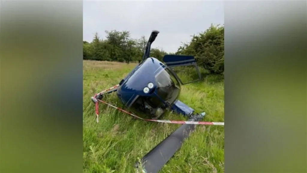 A small blue helicopter lying on its side in a grassy field with its rotor blades damaged.