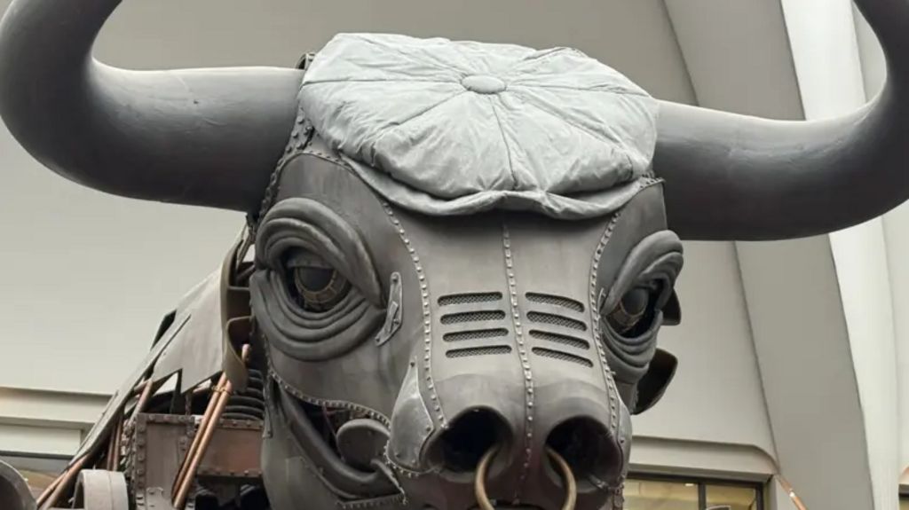 The  head of a mechanical bull, wearing a large grey flat cap. There is a light-coloured wall behind the bull. Its horns and the rest of its body extend out of the shot.