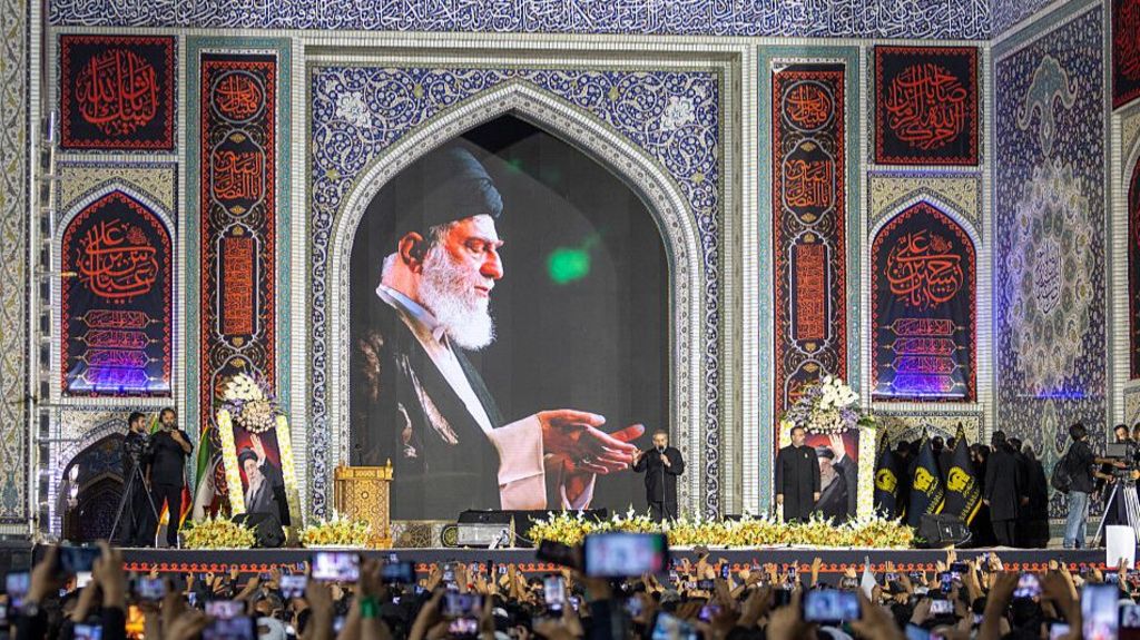 A crowd holds their phones up to take pictures at the funeral of Ayatollah Ali Khamenei, a picture of whom appears above a stage.