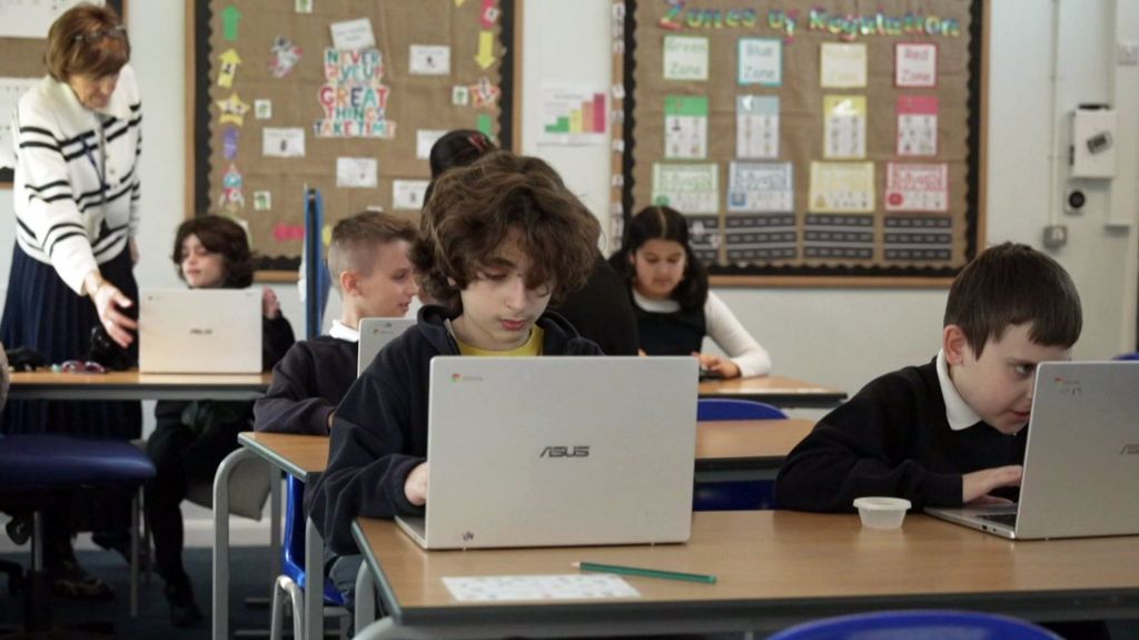 School children sitting at desks in a classroom setting with laptops.