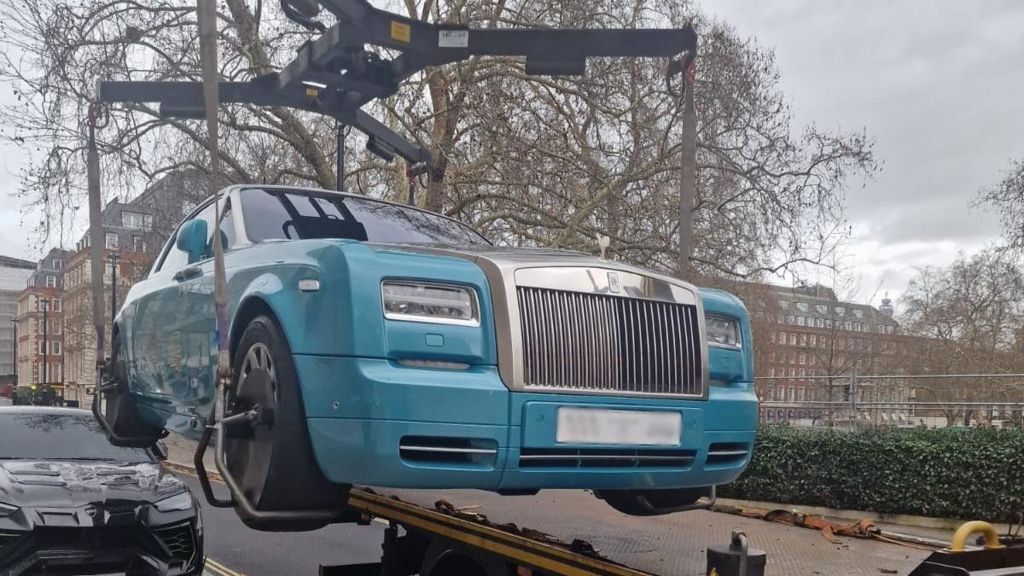A blue Rolls Royce hangs suspended in the air as it is loaded on to a relocation vehicle. 