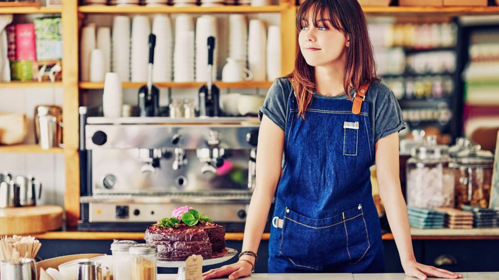 A young woman with long reddish-brown hair and wearing a dark blue apron stands behind the counter in a brightly-lit cafe, next to an ornate chocolate cake. There is a large coffee machine on the back wall, with piles of empty take-away cups and several boxes of tea bags. 