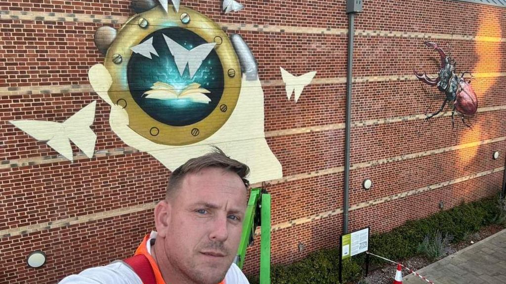 Nathan Murdoch in a selfie-style picture in front of his large scale mural on the back of The Hold archives building in Ipswich. He is pictured looking at the camera while wearing an orange high-viz jacket.