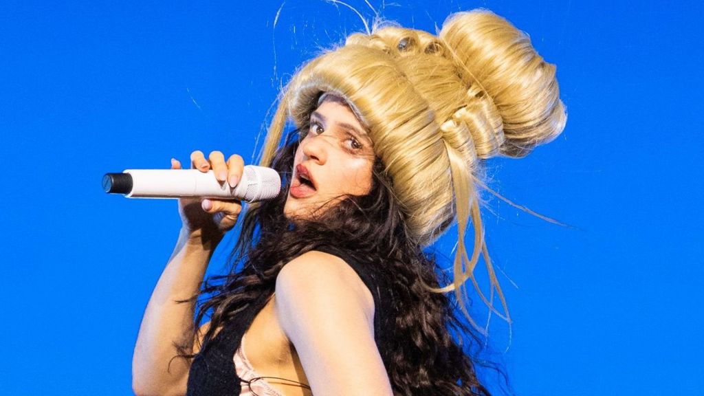 Roslaia on stage wearing a blonde beehive wig and singing into a handheld microphone