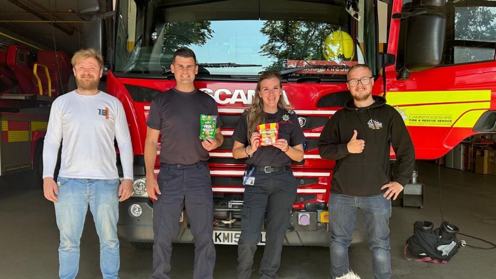 Two firefighters stand in front of a fire engine holding up sweets, with the two men who brought the snack to them 