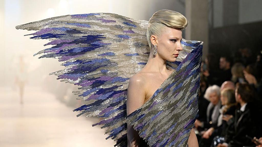 A model with swept back blonde hair in a quiff wearing an elaborate sculptural beaded top, in purples, blues and silvery white, which swoops behind her with the effect of it looking like a wing