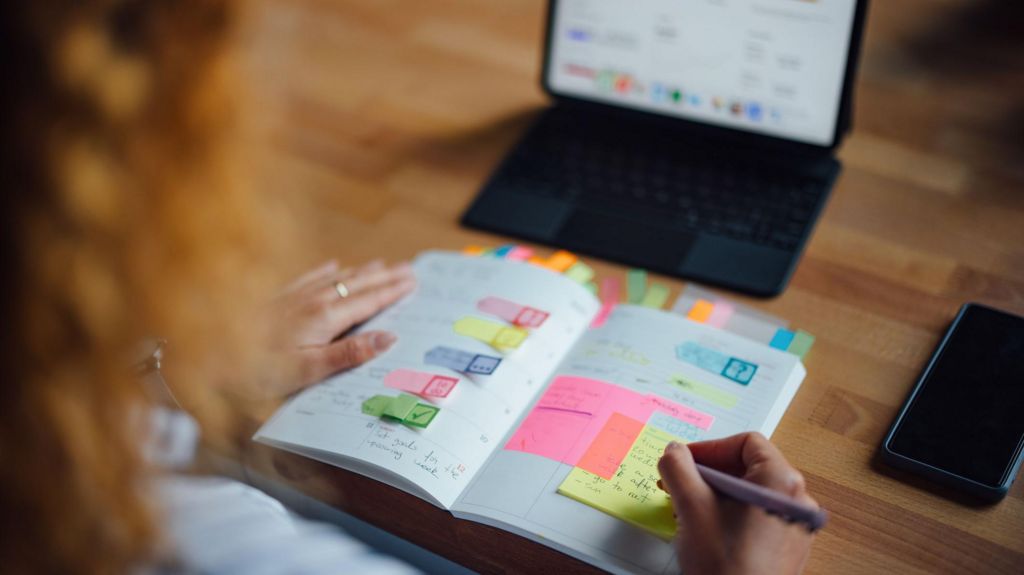 A person with their diary in their hand while looking at a tablet propped up on a table. The diary has lots of colourful tabs and post-its in it