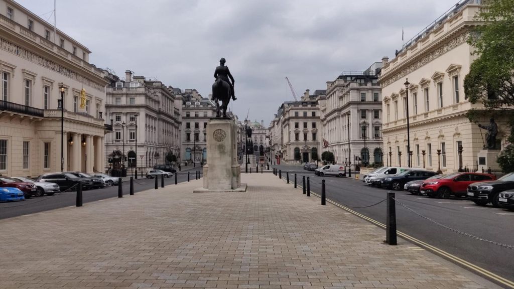 An equestrian statue on a stone plinth stands at the centre of Waterloo Place, with grand historic buildings lining both sides of the broad street in central London.