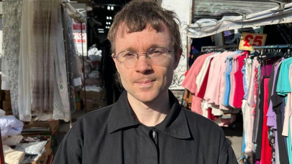 A man with light brown hair and glasses stands between rows of clothes.