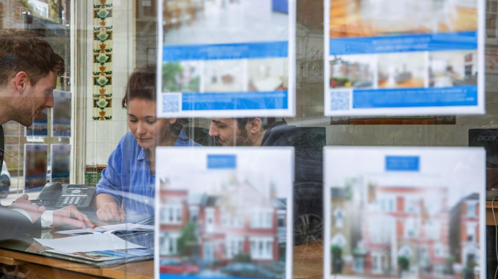 Couple looking at paperwork in an estate agent with pictures of homes for sale in the foreground.