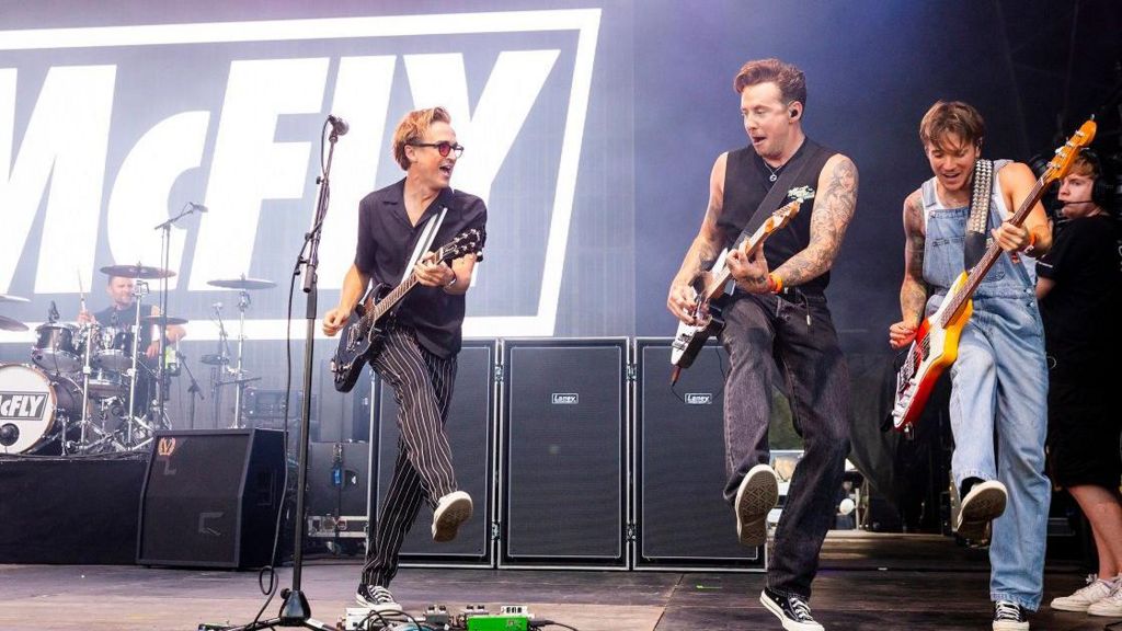 Members of McFly perform on stage. Three men hold guitars and all are kicking their right foot in the air at the same time