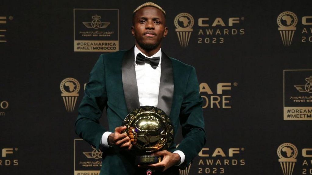 Victor Osimhen in green dinner jacket and black bow tie poses with his African Player of the Year trophy in front of boards promoting the Caf Awards 2023